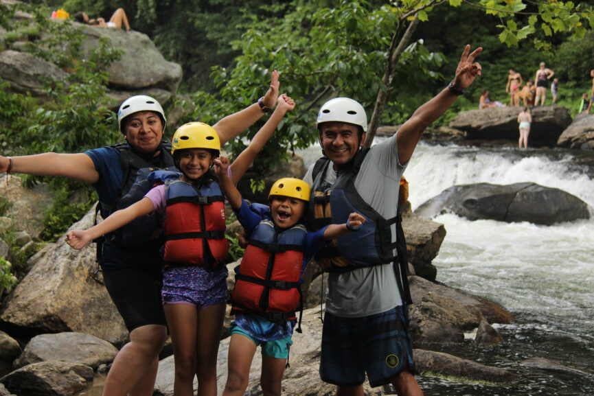 Family of 4 standing in front of the river excitedly with their arms up